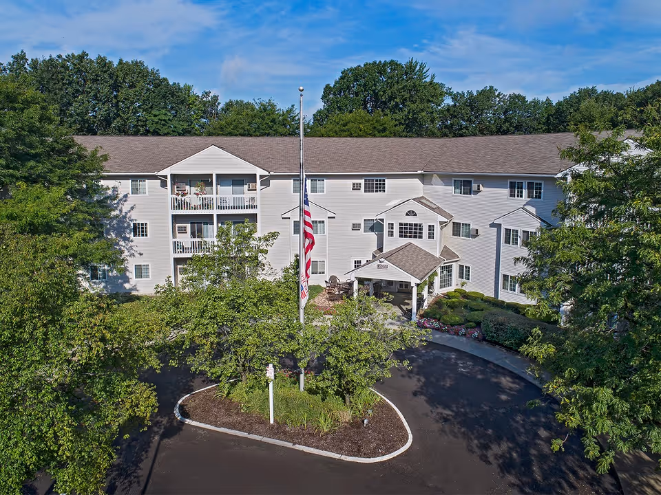 Front exterior of a multi-story senior living building with a covered entrance, flagpole, balconies, and surrounding trees.