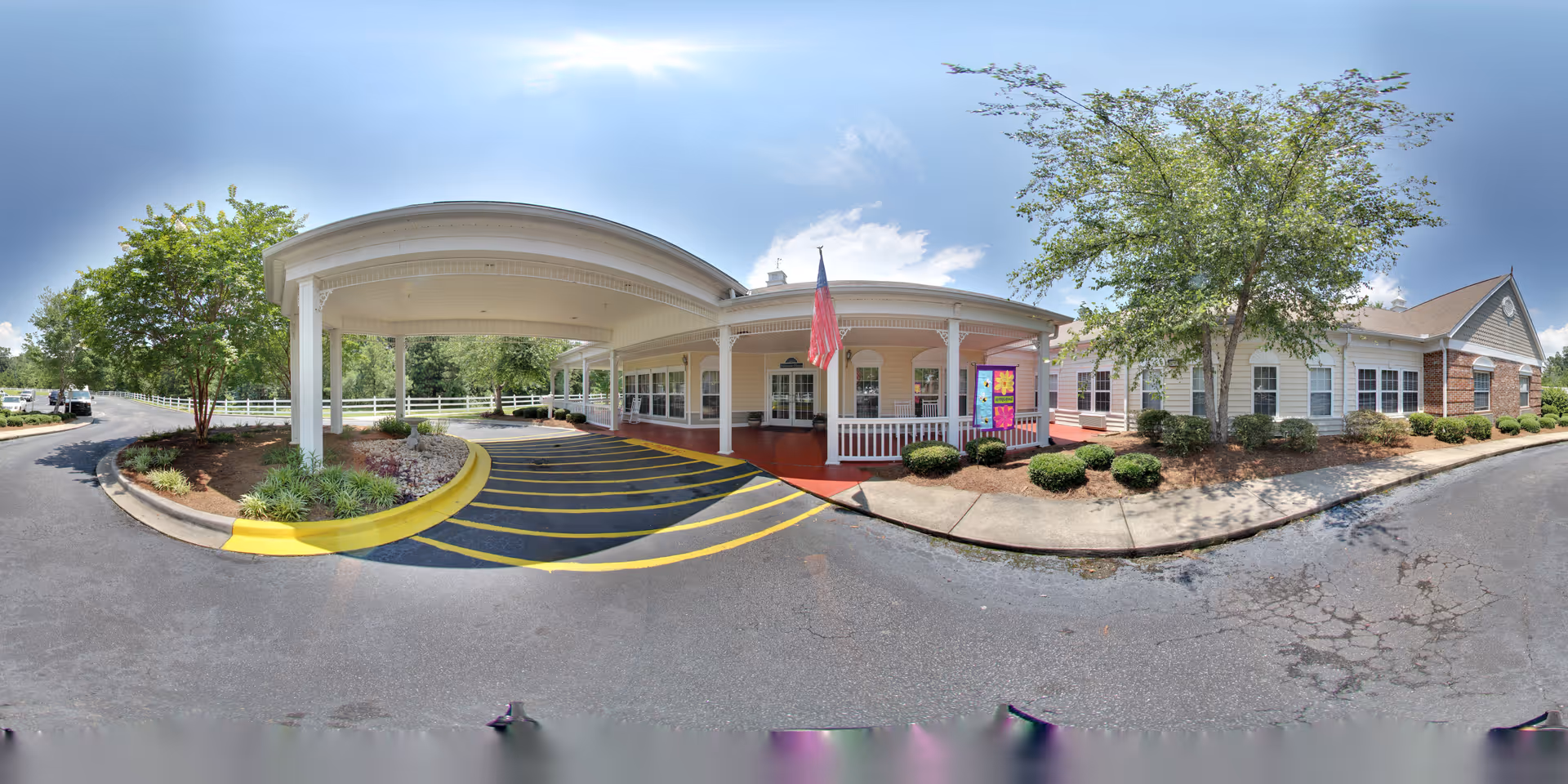Front entrance and covered porte-cochère of a single-story senior living building with an American flag, driveway markings, and surrounding landscaping.