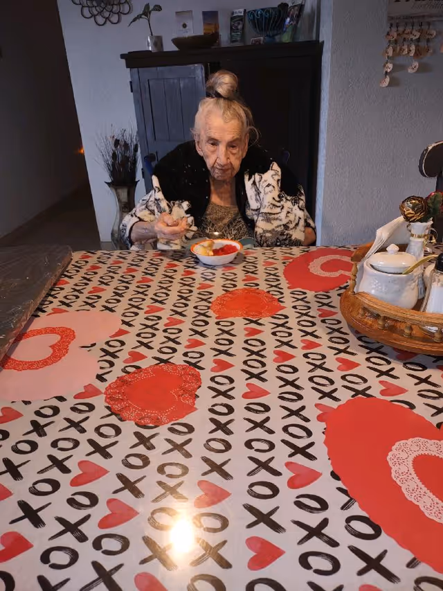 An elderly woman with her hair in a bun sits at a table covered with a tablecloth decorated with red hearts and black XOXO patterns. She is eating from a bowl with a spoon. Behind her is a dark wooden cabinet and some wall decorations.