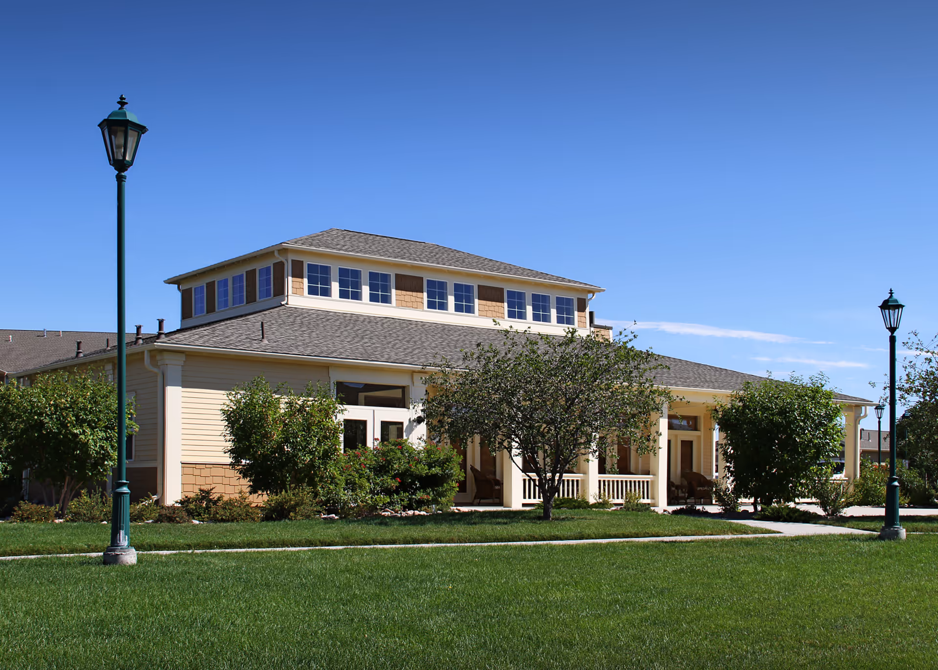 Exterior view of a single-story building with a small upper section featuring multiple windows. The building is surrounded by green grass, bushes, and trees, with two vintage-style street lamps on either side. The sky is clear and blue.