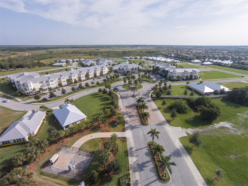 Aerial view of The Brennity at Tradition Senior Living facility showing multiple buildings with light-colored roofs, surrounded by green lawns, palm trees, and roads. The layout includes a circular driveway and parking areas, with open fields and a distant residential area in the background under a partly cloudy sky.