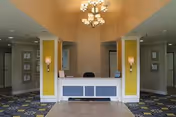 A bright hotel-style lobby with yellow walls, a white-and-blue reception desk, and chandeliers.