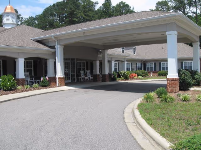 Entrance area of a senior living facility with a covered driveway supported by white columns with brick bases. The building has a gray roof and beige siding, with several windows and rocking chairs on the porch. There are landscaped bushes and plants along the building and driveway.