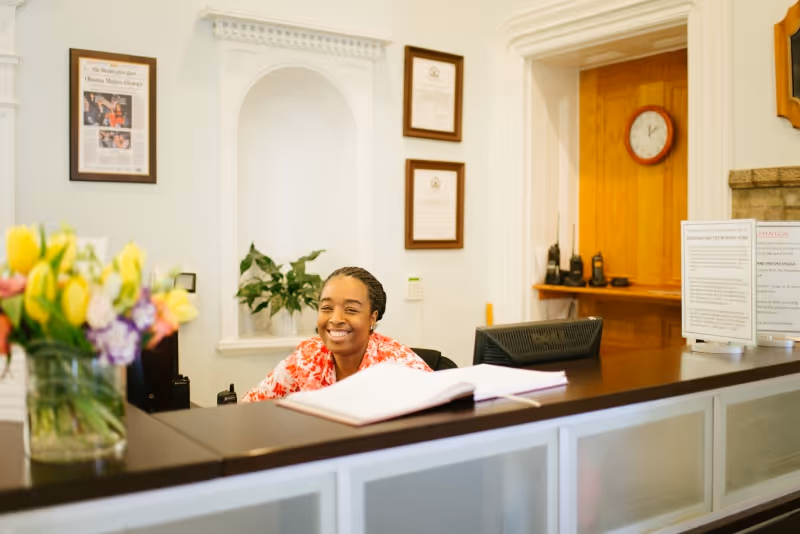 A smiling receptionist sits behind a nursing home reception desk with a vase of flowers, an open guestbook, and framed certificates on the wall.