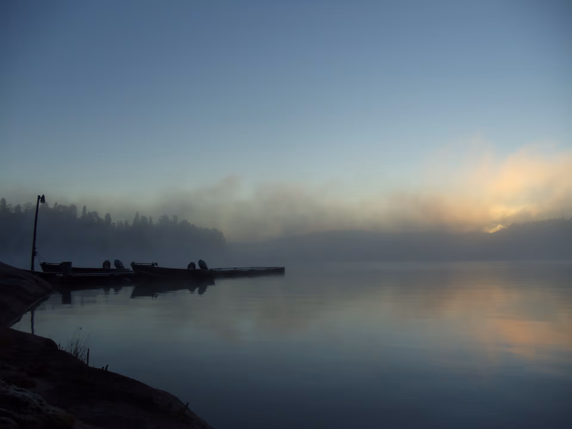 Calm lake at dawn with fog and boats docked along a misty shoreline.
