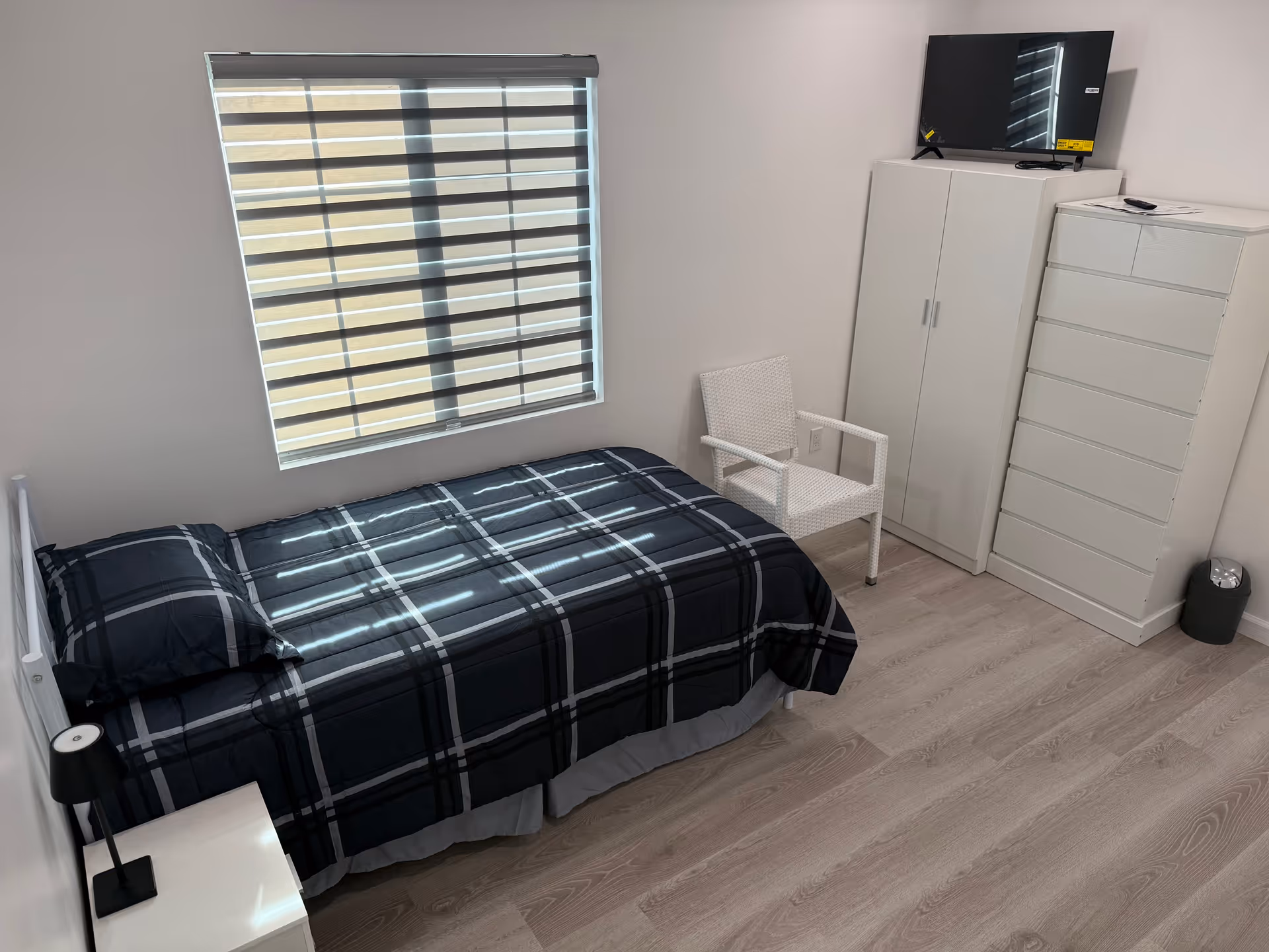 Bedroom with a single bed, plaid bedding, blinds, chair, dresser, and a TV.