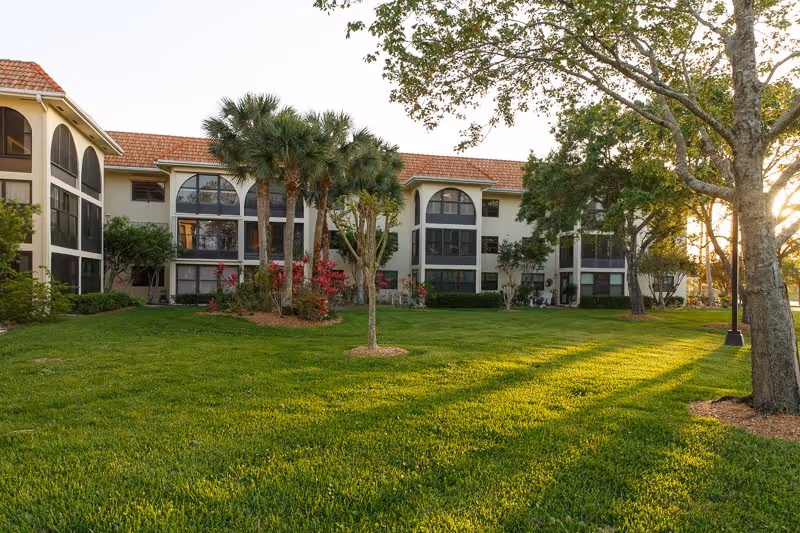 Sunlit exterior of a multi-story senior living building with arched windows, palm trees and a manicured lawn.