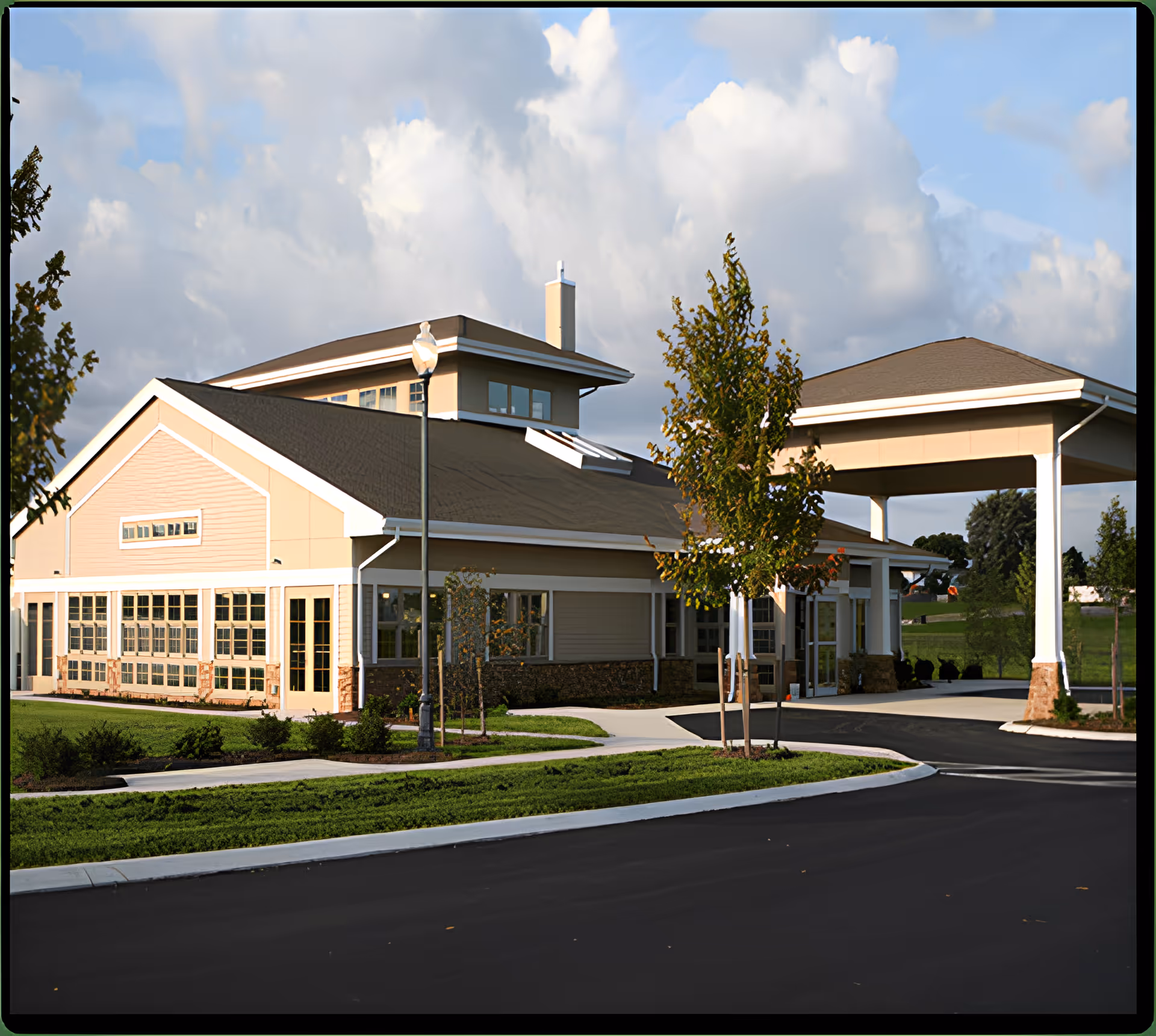Front exterior of a single-story senior living building with a covered entrance, large windows, and landscaped grounds under a cloudy sky.