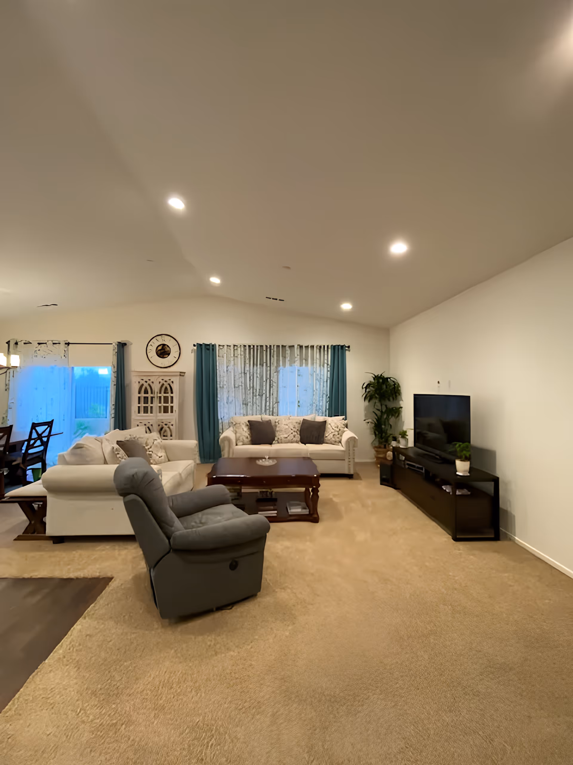 A spacious living room with beige carpet and white walls, featuring two white sofas with patterned cushions, a gray recliner chair, a wooden coffee table, a TV on a black stand, and a tall green plant. The room has recessed ceiling lights and windows with blue curtains and patterned sheer curtains.