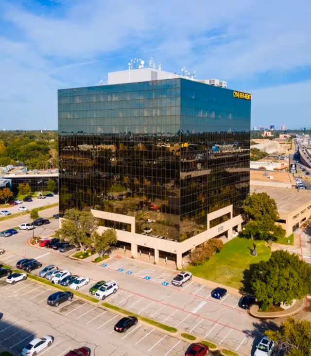 Glass-clad office building with parking lot, trees, and a blue sky overhead.