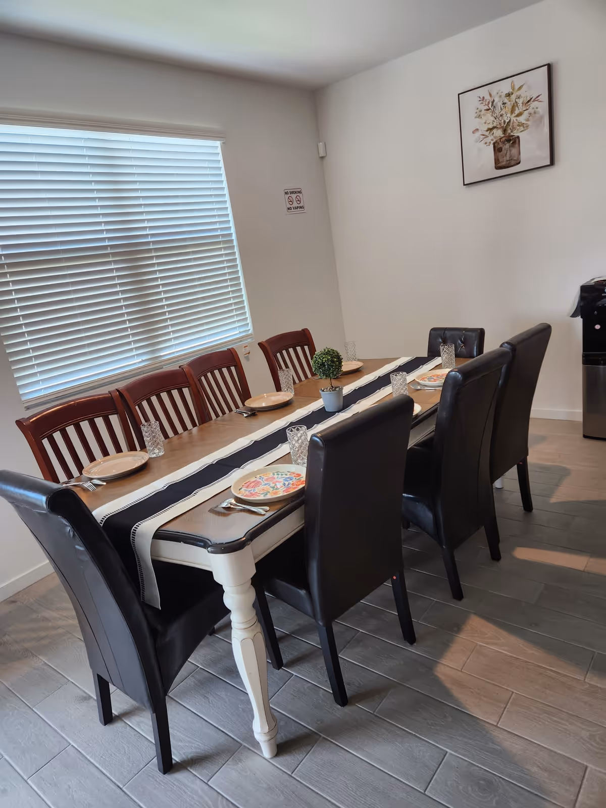 A dining room with a long wooden table set for six people. The table has a white and black runner, six plates with cutlery, and six glasses. There are three black cushioned chairs on one side and three wooden chairs on the other. A small potted plant is placed in the center of the table. The room has a window with closed blinds, a 'No Smoking No Vaping' sign on the wall, and a framed floral painting hanging on a white wall.