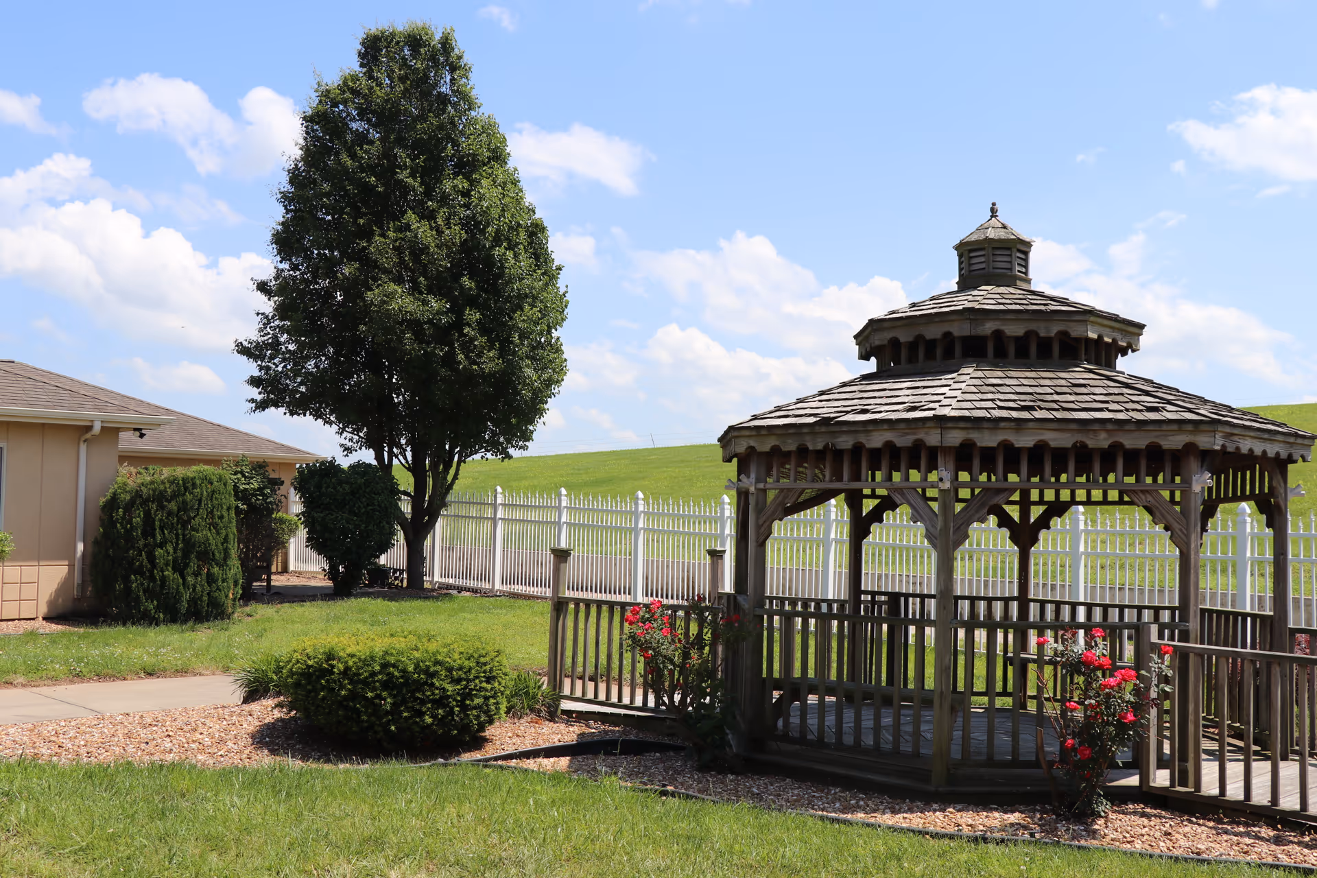 Outdoor area of Ash Grove Healthcare Facility & Rehabilitation featuring a wooden gazebo surrounded by green grass, bushes, and a tree, with a white fence and a building partially visible in the background under a blue sky with scattered clouds.