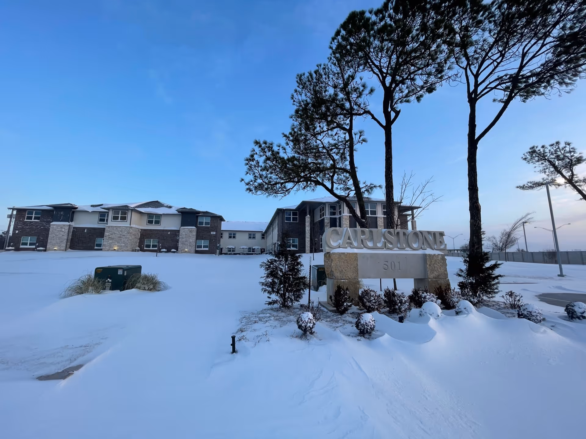Snow blankets Carlstone Active Adult Living Community's front grounds, featuring a large stone sign and trees.