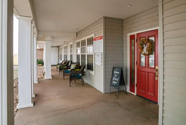 Covered porch area with white columns and a red door decorated with a wreath. Several chairs with cushions are arranged along the wall with windows. A small chalkboard sign near the door reads 'Welcome to Hickory Terrace'.
