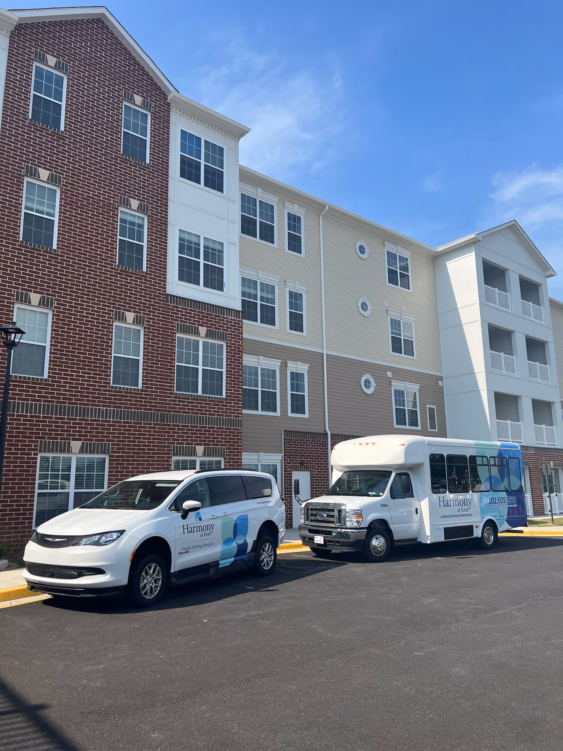 Exterior view of a senior living facility named Harmony at Kent showing a multi-story building with brick and siding facade. Two white vehicles with Harmony at Kent branding are parked in front, including a van and a shuttle bus. The sky is clear and blue.