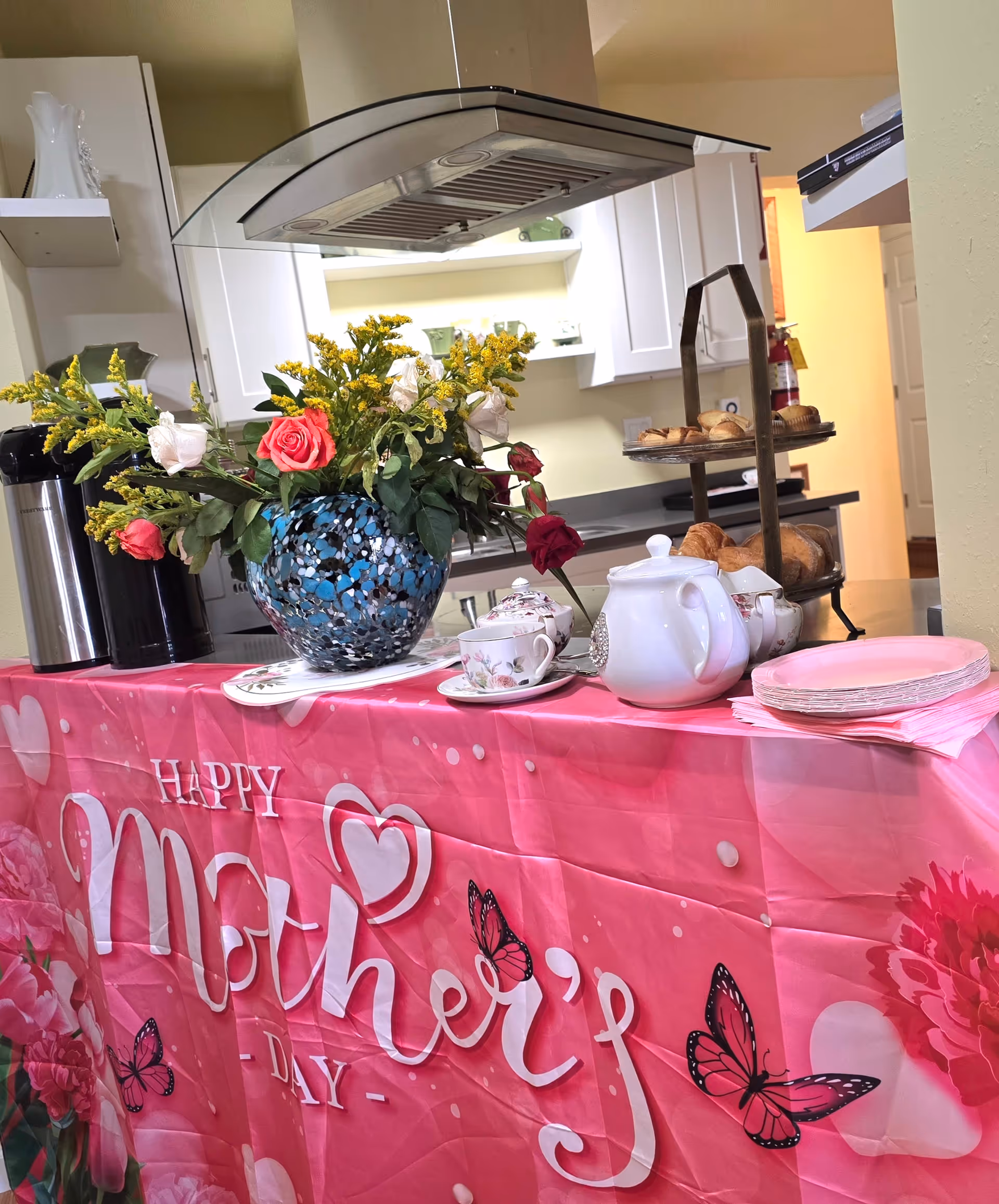 Pink Mother's Day tablecloth on a counter with flowers, teapot, cups, and pastries in a kitchen.