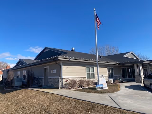 Single-story beige senior living facility building with a flagpole and curved driveway under a clear blue sky.