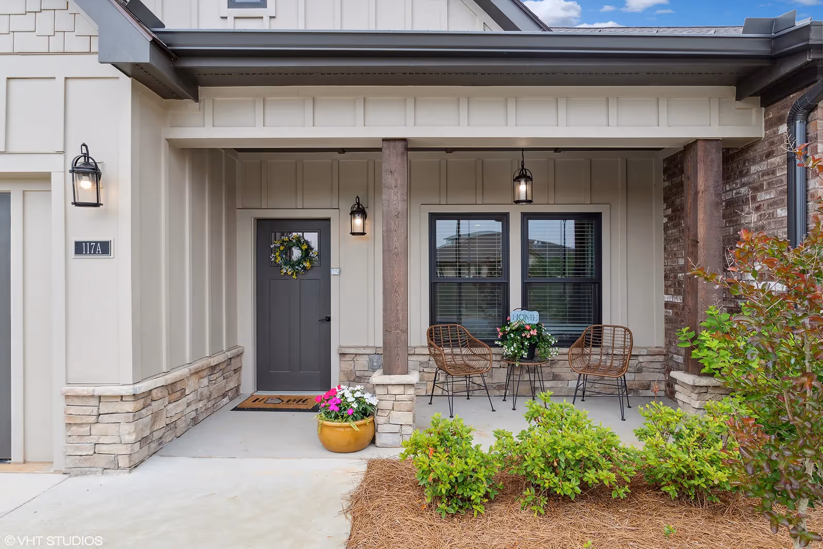 Front porch of a residential unit with a gray door decorated with a wreath, two wicker chairs and a small table with a flower pot and a 'HOME' sign between them, stone and wood exterior accents, and some green shrubs in front.