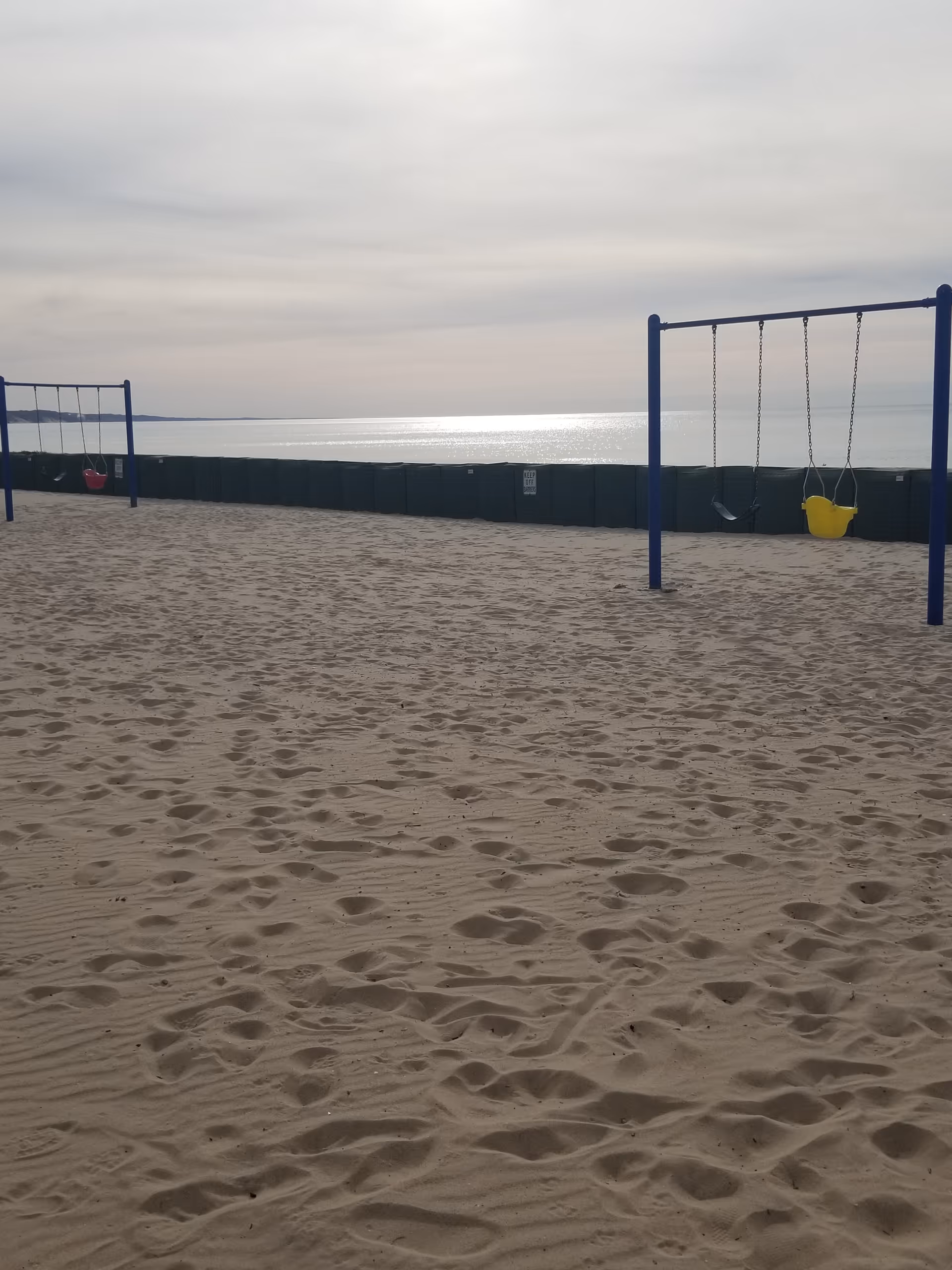Sandy beach with blue swings and calm water on the horizon under overcast sky.