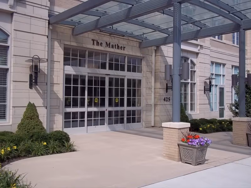 Exterior view of a senior living facility entrance with glass doors, plants, and a covered walkway.