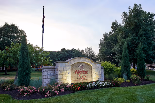 Stone entrance sign reading "Wexford Place" surrounded by landscaped flowers, trees, and a flagpole at a senior living community.