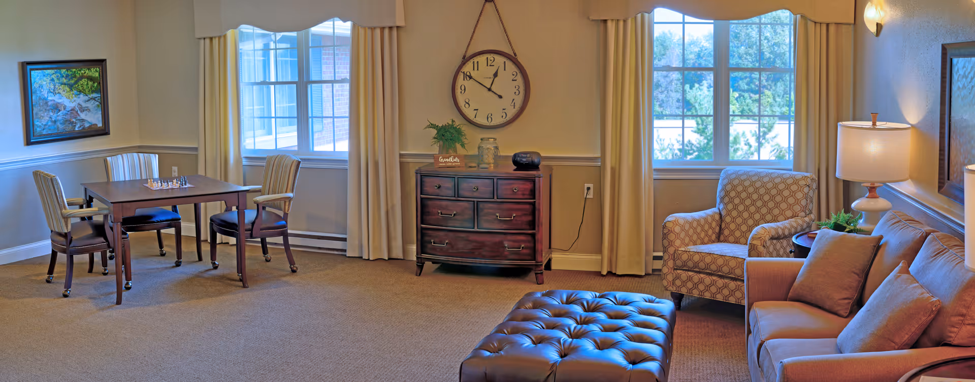 A cozy living room area with a beige sofa, an armchair, a tufted ottoman, and a wooden side table with a lamp. There is a wooden chest of drawers against the wall with a round clock hanging above it. Two large windows with beige curtains let in natural light. A small table with four chairs and a chessboard is positioned near one window. The walls are decorated with a framed painting and a wall sconce.