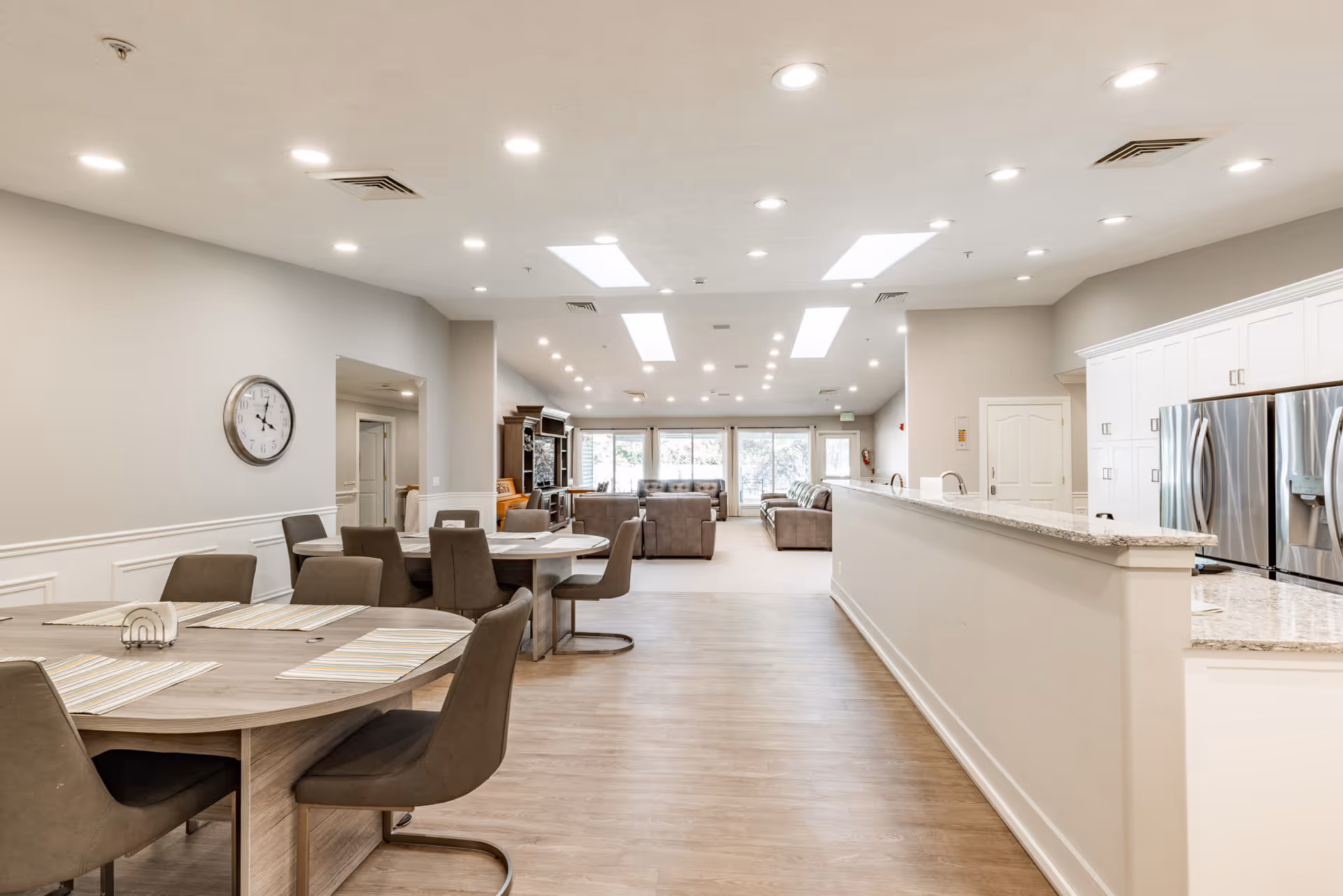 Open, well-lit senior living common area with round dining tables and a granite kitchen counter.