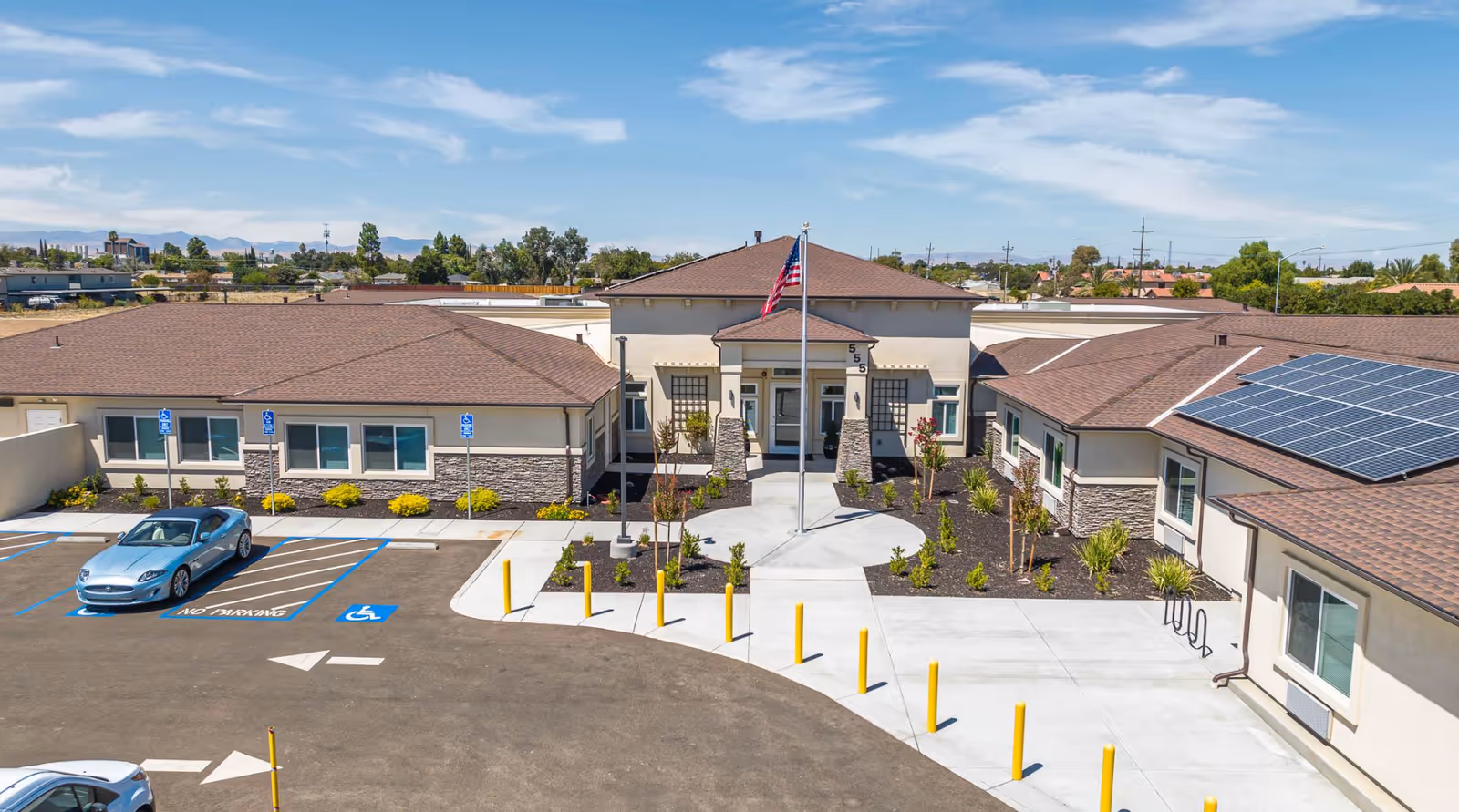 Entrance and parking lot of a single-story memory care facility with an American flag, handicap parking spaces, and solar panels on the roof.