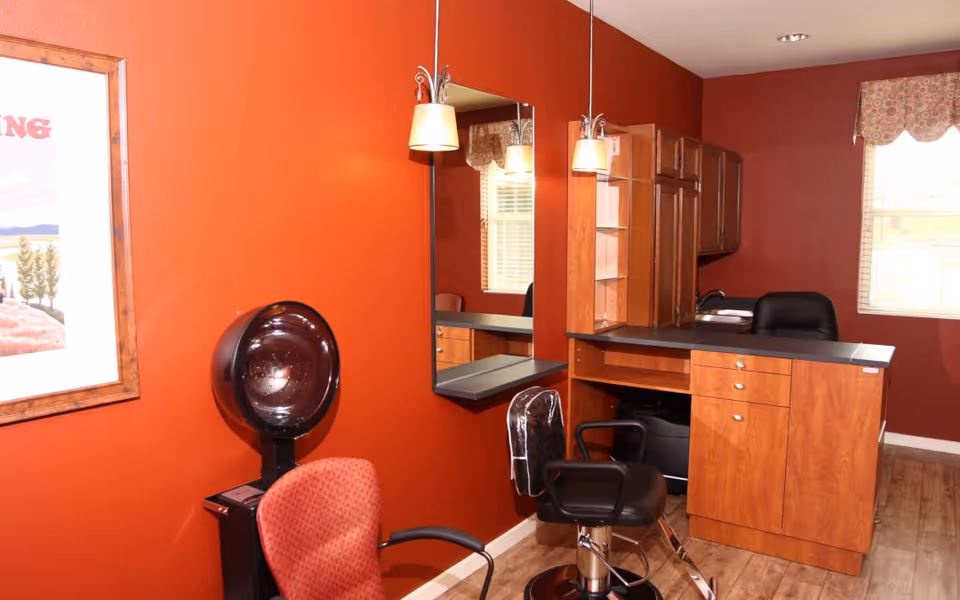 Salon-style room with red walls containing a styling chair, hooded hair dryer, wall mirror, and wooden cabinets and counter.