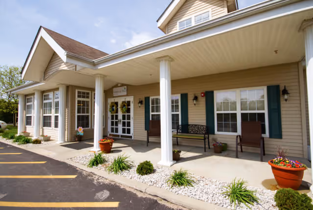 Exterior view of Whitlock Place senior living facility showing a beige building with white columns supporting a covered porch. There are potted plants, benches, and chairs on the porch, with a clear blue sky above.