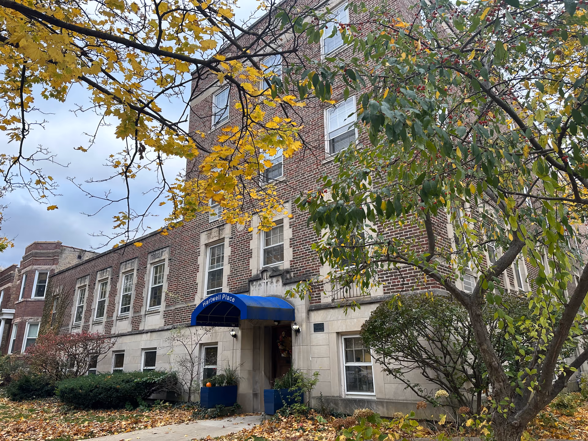 Exterior view of a multi-story brick building with a blue awning labeled 'Hartwell Place' over the entrance. The building is surrounded by trees with green and yellow leaves, and fallen leaves cover the ground, indicating an autumn setting.