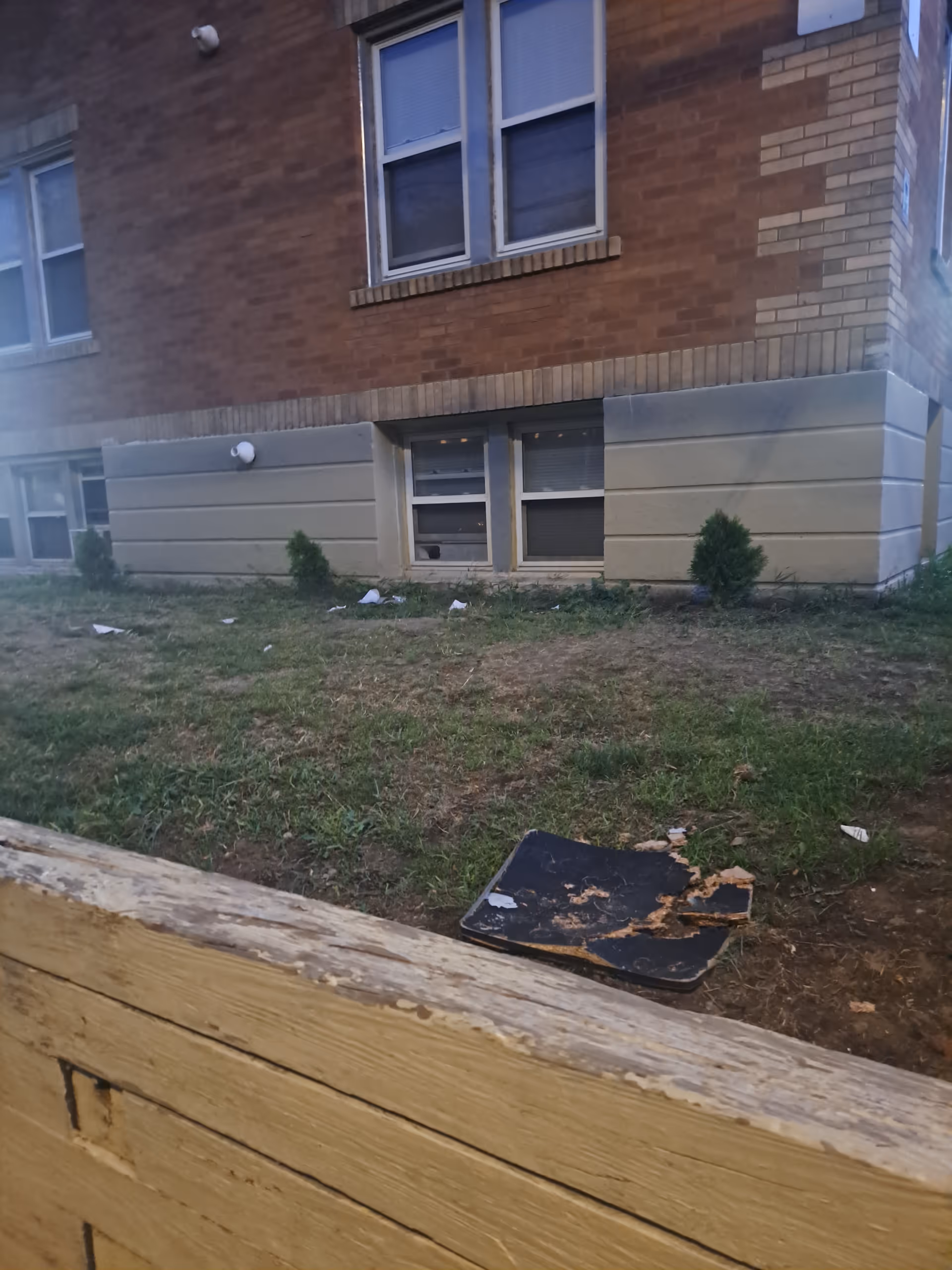 Exterior brick building with a grassy yard, scattered debris, and a wooden railing in foreground.