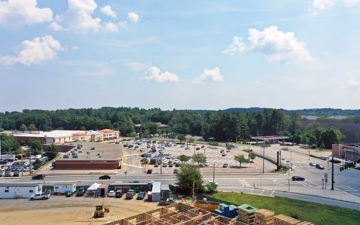 Aerial view of a large parking lot with multiple cars parked, surrounded by commercial buildings and a road with vehicles. In the foreground, there is a construction site with wooden framing and construction equipment. Trees and a partly cloudy sky are visible in the background.