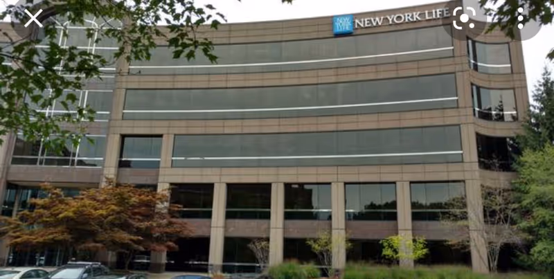 Modern glass-front office building with trees, parked cars, and a New York Life sign at top.