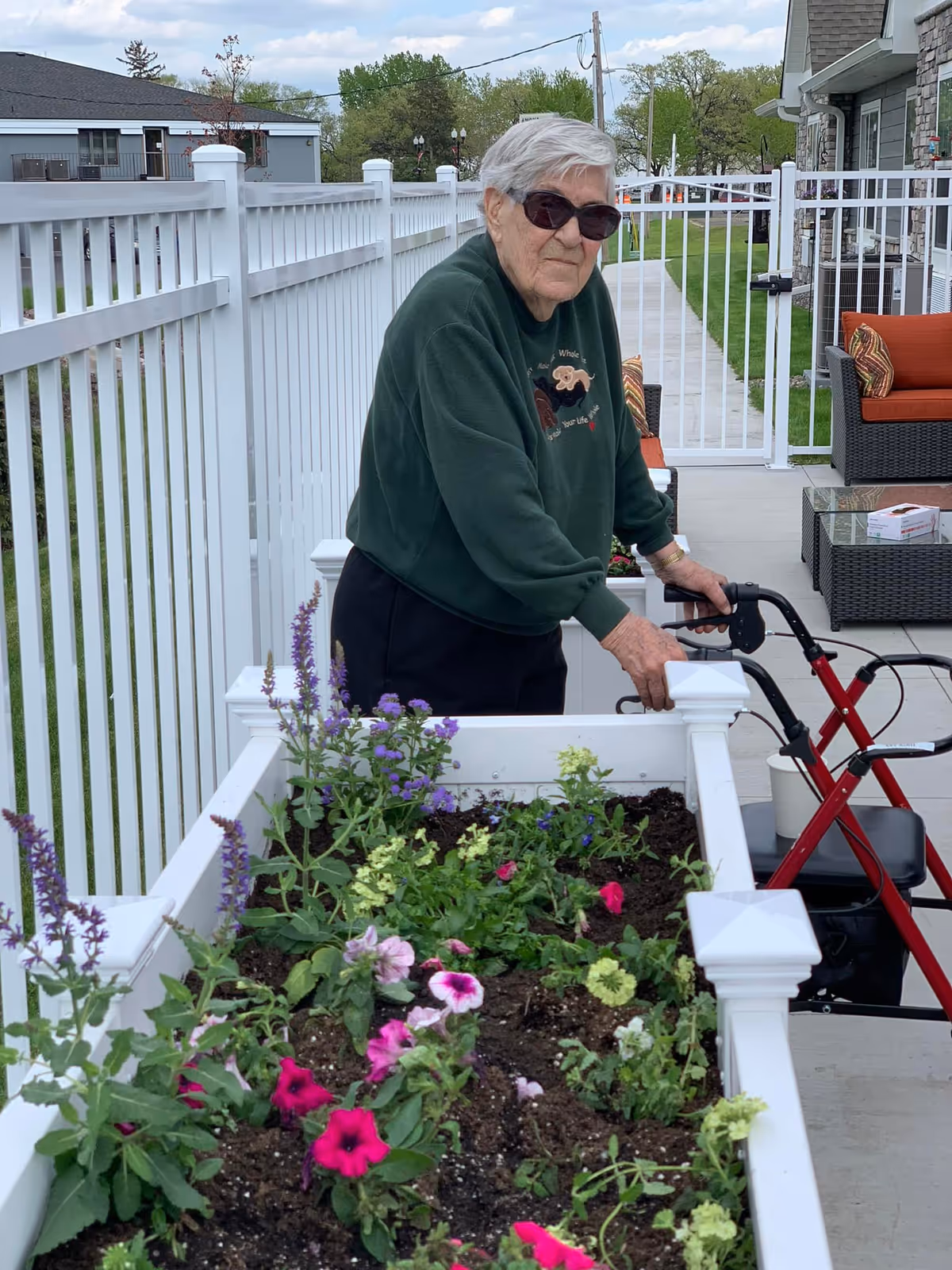 An elderly person using a walker, tending to flowers in a raised garden bed outside a senior living facility.