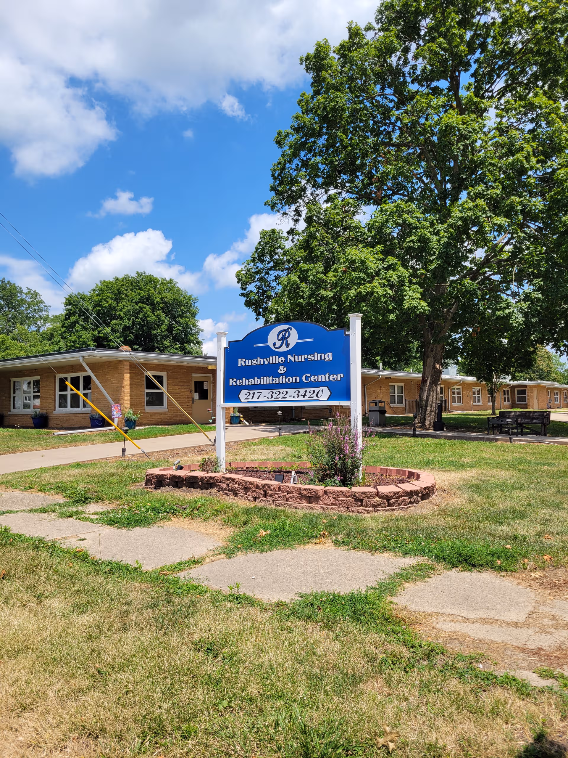 Blue Rushville Nursing and Rehabilitation Center sign in front of a low brick building with trees.