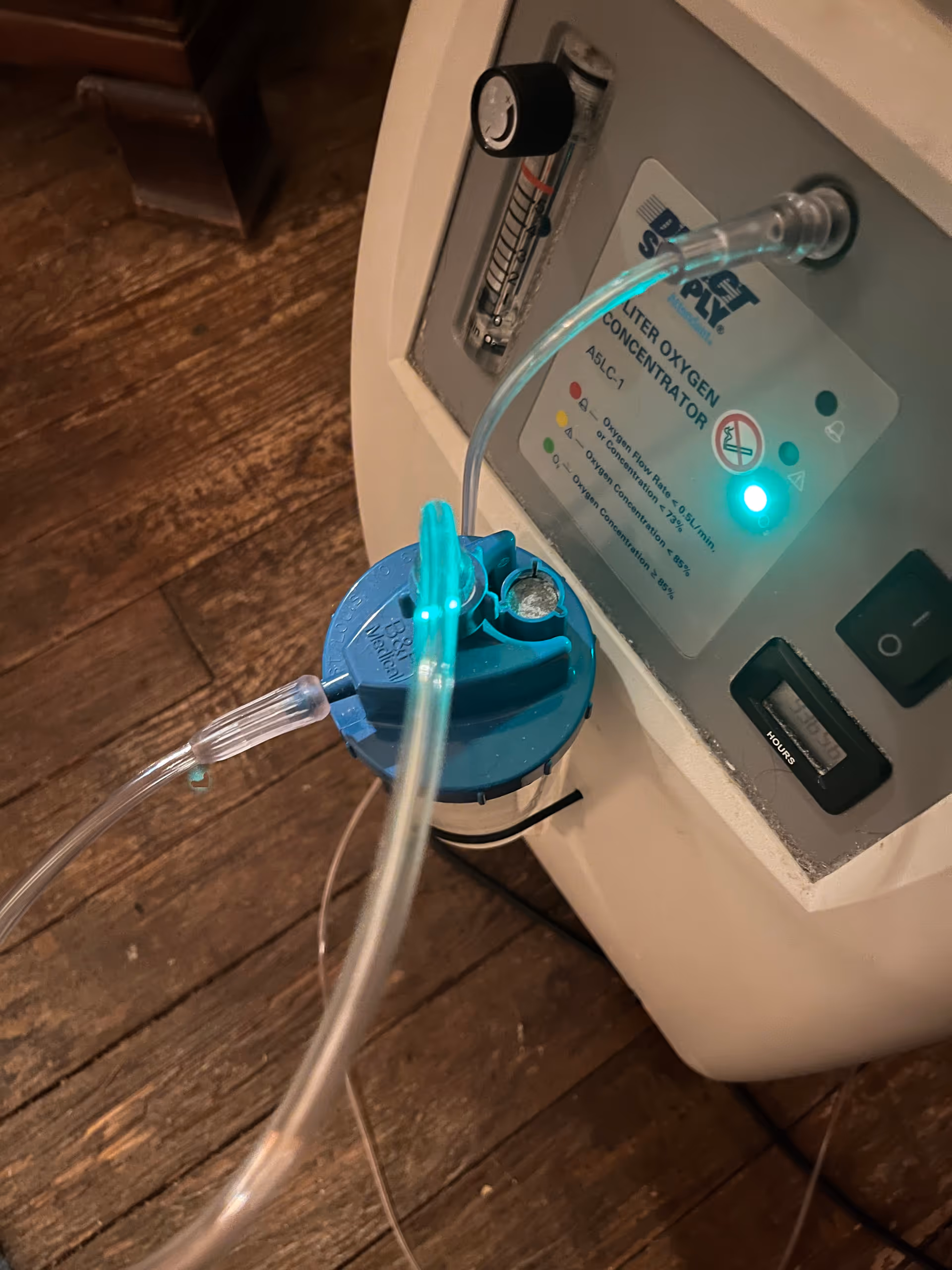 Blue oxygen concentrator with tubes sits on a wooden floor indoors.
