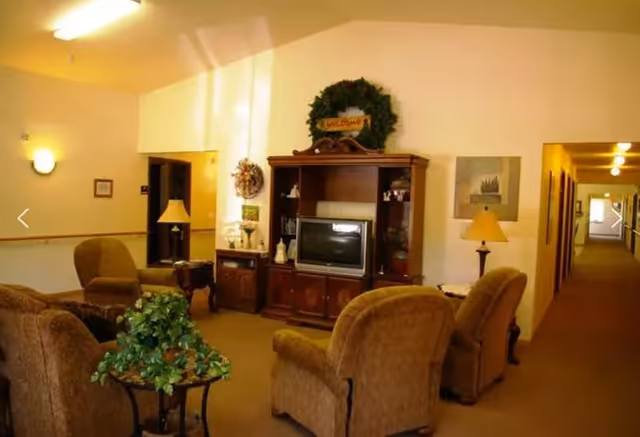 A cozy living room area with four upholstered armchairs arranged around a small table with a green leafy plant. Against the wall is a wooden entertainment center with a television and decorative items, topped with a green wreath and a 'Welcome' sign. The room is warmly lit with lamps and wall sconces, and a hallway extends to the right.