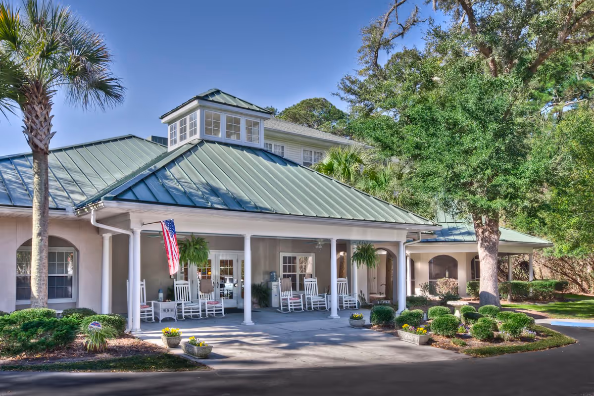 Exterior view of The Pines at Bluffton Assisted Living & Memory Care Community building with a green metal roof, white columns, rocking chairs on the porch, an American flag, and surrounding greenery including palm trees and bushes.