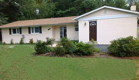 Single-story light-colored building with a peaked roof, brown front door, windows, and a grassy lawn with shrubs.