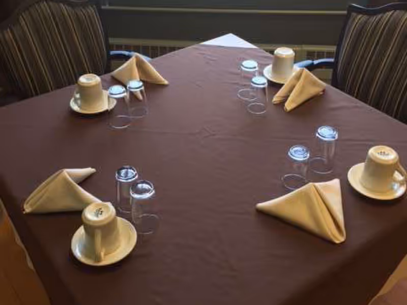 Round dining table with brown tablecloth, folded napkins, cups, and glasses in a dining room.