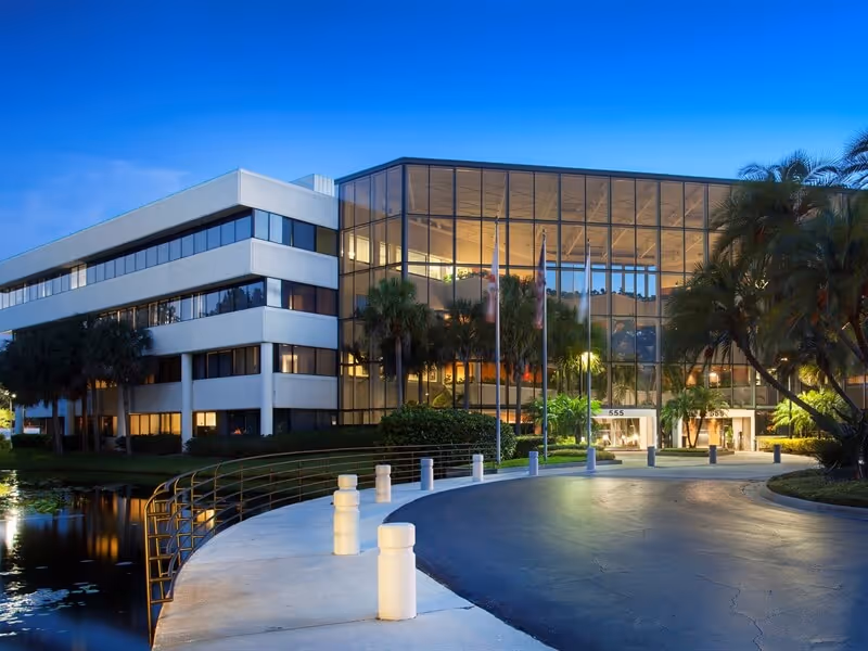 Modern glass-front senior care facility with palm trees, curved driveway, and dusk reflections.