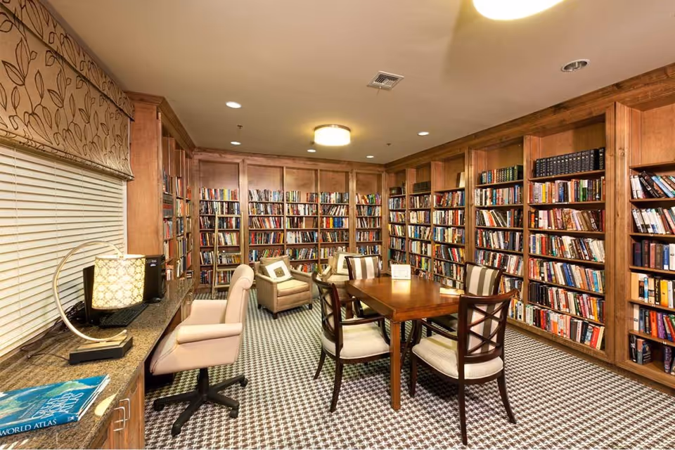 A cozy library room with wooden bookshelves filled with books lining the walls. In the center, there is a wooden table surrounded by four cushioned chairs. To the left, there is a countertop with a computer and a lamp, along with a comfortable office chair. The room has a patterned carpet and a window with a floral valance.