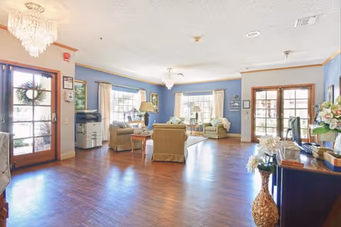 Bright, open senior living common room with seating groups, chandeliers, large windows, and wood floors near glass entry doors.