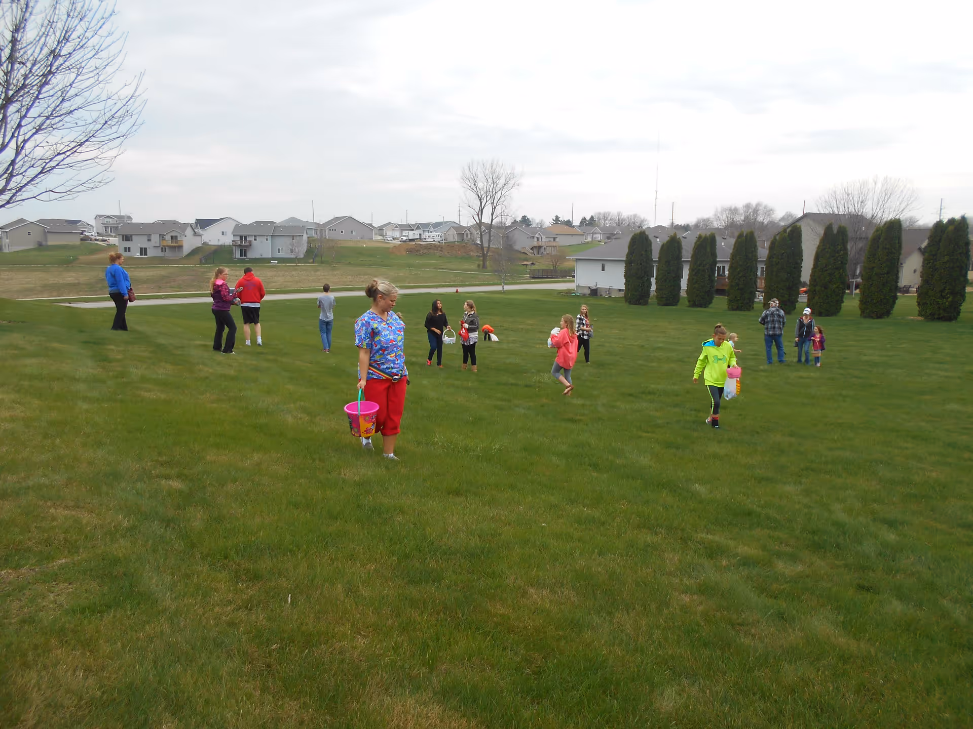A group of people, including children and adults, are scattered across a large grassy field near a residential area. Some children are holding colorful buckets, possibly participating in an outdoor activity or event. The sky is overcast, and there are houses and tall trees in the background.