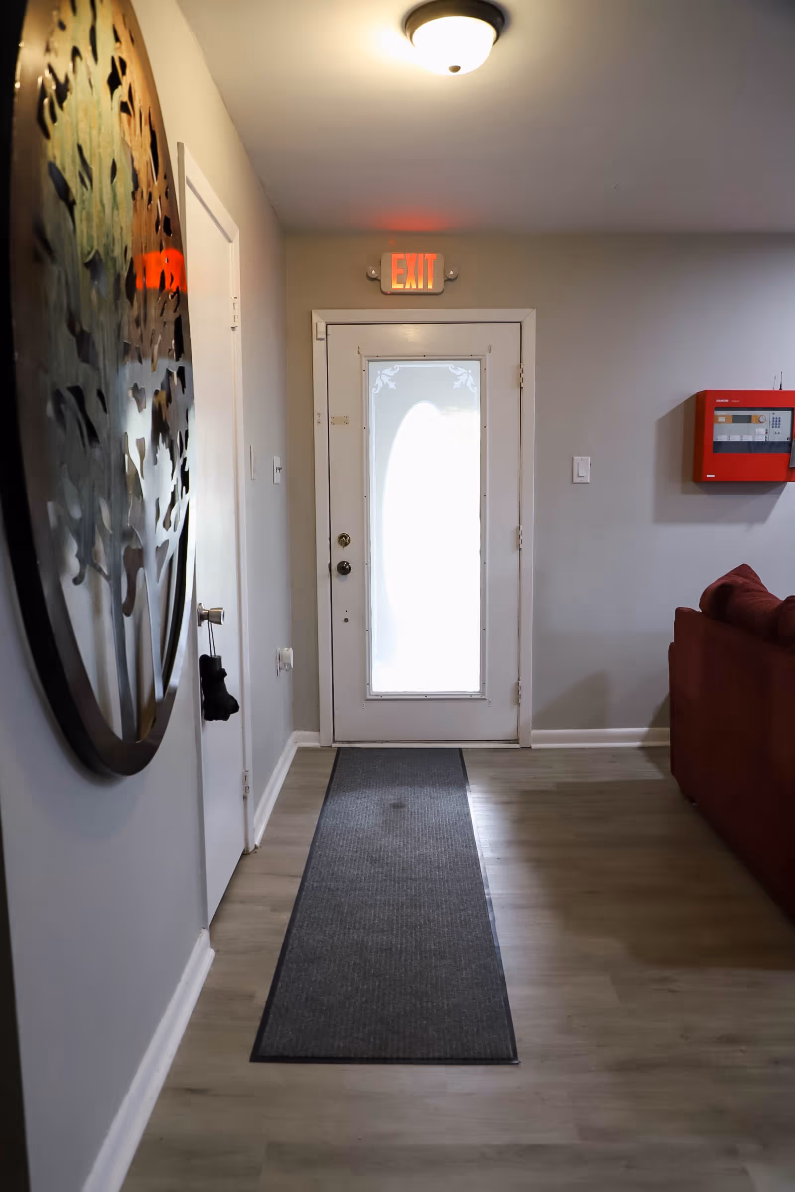 Interior hallway of a facility with a white door featuring a frosted glass oval window, an illuminated red EXIT sign above the door, a gray runner rug on light wood flooring, a large decorative metal wall art on the left wall, a red fire alarm panel on the right wall, and part of a red couch visible on the right side.