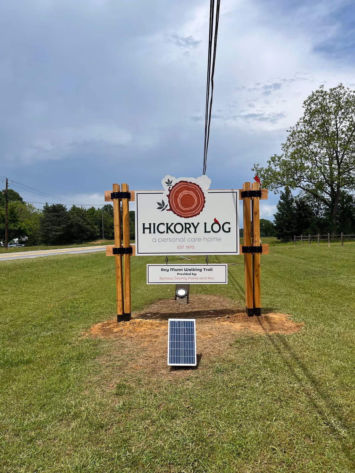 Wood-framed Hickory Log sign on a grassy lawn with trees and a cloudy sky.