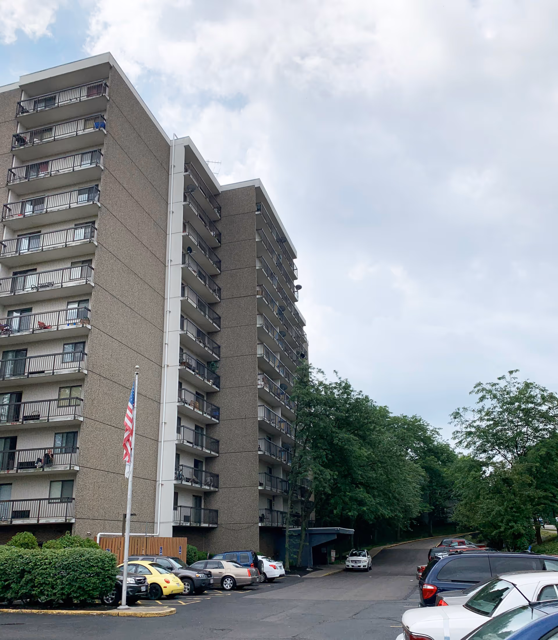 Multi-story residential building with balconies, a flagpole, and parked cars in the foreground under a cloudy sky.