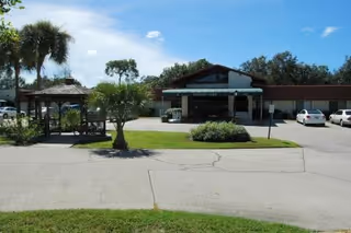 Front exterior of a single-story senior living facility with a covered entrance, gazebo, palm trees, and parked cars.