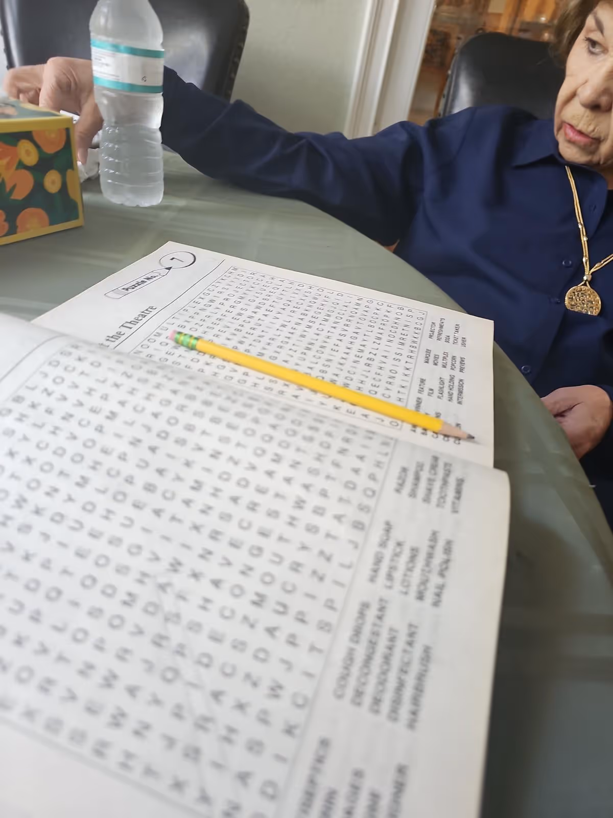 Elderly resident sits at a table solving a word puzzle with a pencil.